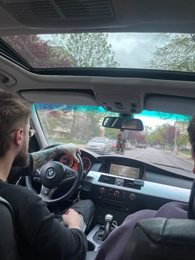The image depicts the interior of a car, specifically a BMW E60/E61 model, as viewed from the front passenger seat. The scene is set in Nova Zagora, Bulgaria, during daytime under an overcast sky.
The primary subject is the male driver, seen from a slightly elevated angle. He has a beard and wears glasses, and his left arm, holding the BMW-branded steering wheel (which also features an M badge), is extensively tattooed. He is wearing a dark, long-sleeved garment.
The car's dashboard is prominent, featuring an infotainment screen displaying menu options like "Navigation," "Communication," "Entertainment," and "Climate," alongside climate control dials, air vents, and a CD/DVD slot. The iDrive controller is visible near the gear shifter. An "areon X" air freshener with a kiss mark design hangs from the rearview mirror.
Through the windshield and the open sunroof, an outdoor street scene is visible. Trees with both green and reddish-brown leaves line the street, suggesting either a spring or autumn setting. Other vehicles can be seen on the road. The sky is cloudy, indicating an overcast day. A person's head and shoulder are partially visible in the upper right portion of the image (when oriented correctly), likely the individual capturing the photograph. The overall atmosphere suggests a casual drive through an urban or suburban environment.