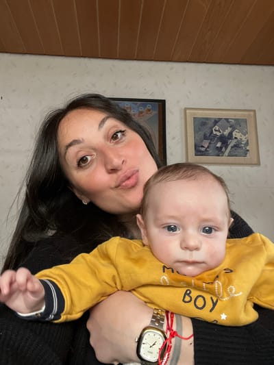 This image depicts an adult female holding an infant indoors in Wüstenrot, Germany. The female, positioned centrally, has dark hair and brown eyes, and is making a pouting expression towards the camera. She wears a dark garment, small stud earrings, a silver-toned rectangular wristwatch, and red string bracelets on her left wrist. The infant, cradled by the female, has light hair and blue eyes, and wears a yellow long-sleeved shirt with the partial inscription "LITTLE BOY" and a white star. The infant maintains a neutral gaze towards the camera. The background wall features light-colored, textured wallpaper. Above, a wooden-paneled ceiling is visible. Two framed pictures are mounted on the wall: the left frame is dark, containing an image of stylized figures; the right frame is light wood, displaying an aerial view of residential structures.