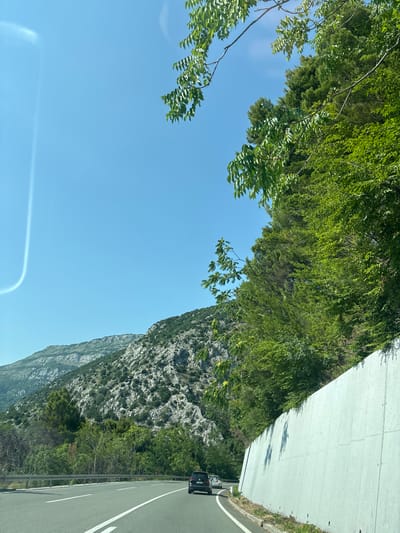 This media file captures a scenic view from a vehicle traveling along a road, likely in a mountainous region. The scene is dominated by a clear blue sky, lush green foliage, and a rugged, rocky mountainside. In the foreground, branches and leaves from trees hang into the frame, obscuring parts of the sky. The road itself is visible on the right side of the image, marked with white lines, and a black vehicle is seen ahead, traveling in the same direction. A concrete barrier separates the road from the embankment which is covered in dense vegetation. The time of day appears to be daytime, with bright sunlight casting shadows. The overall impression is one of a picturesque journey through a natural landscape.