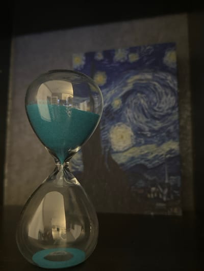 The media file displays a close-up, horizontal shot of an hourglass with teal-colored sand. The hourglass is positioned in the foreground, with its glass bulbs reflecting ambient light and what appears to be an indoor scene with furniture. In the background, slightly out of focus, is a reproduction of Vincent van Gogh's painting, "The Starry Night." The painting features a swirling, dark blue sky filled with bright yellow stars and a crescent moon, with a dark cypress tree dominating the right side and a village nestled in the distance. The overall lighting suggests an indoor setting, likely during the day given the reflections in the glass, but without definitive cues for time of day or weather. No text is visible in the image. The scene's context is given as Hamburg, Germany, but this cannot be visually confirmed from the provided image. The arrangement of the hourglass and the artwork implies a decorative or artistic display.