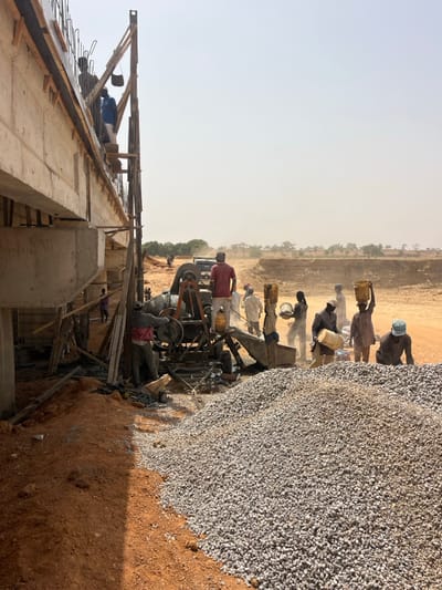The image depicts an active construction site in the city of Gyellesu, Nigeria, under bright, hazy daylight conditions. A concrete bridge or overpass structure occupies the left side, with visible formwork and rebar. Several workers are present:
* Two individuals are positioned on wooden scaffolding attached to the underside of the bridge structure on the left, one carrying a dark bucket. Another worker is partially visible further back under the bridge.
* In the center, a portable concrete mixer is operational. One worker in a red shirt stands on the mixer's loading ramp, while another in a grey jacket operates its controls.
* Around the mixer and extending to the right, approximately eight additional workers are engaged in material handling. Several are carrying yellow or white plastic buckets, some balanced on their heads or shoulders, indicating manual transport of aggregates or mixed concrete. One worker is pouring contents from a white bucket.
* In the immediate foreground, a substantial pile of grey aggregate (crushed stone/gravel) rests on the reddish-brown earth. The ground is unpaved, dusty, and uneven.
* In the middle ground, a light-colored pickup truck is partially visible near the concrete mixer. The background shows an arid landscape with distant scrub brush and trees, and expansive areas of excavated reddish-brown earth, characteristic of large-scale ground disturbance. Dust is visible in the air.