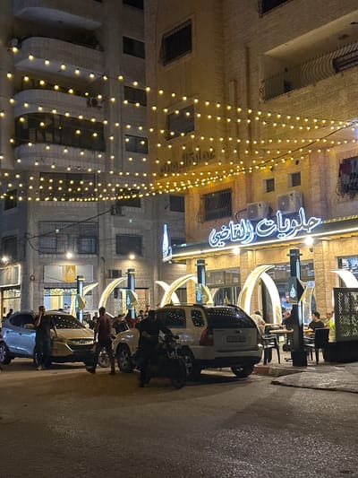 This nighttime exterior photograph captures a street scene in Gaza City, Palestinian Territories. The foreground features a paved road with several cars parked and people standing around them. A light-colored SUV is prominently positioned in the center, with a motorcycle to its right. Several individuals, dressed in dark clothing, are visible near the vehicles.

In the background, a brightly lit building with an illuminated sign in Arabic script stands out. The sign appears to be for a business, possibly a restaurant or sweet shop, indicated by the word "حلويات" (sweets) and "القاضي" (Al-Qadi). The building's facade is made of light-colored stone. Strings of small, yellow lights are strung across the street, illuminating the area and adding a festive ambiance. The architecture of the surrounding buildings suggests an urban environment with multiple stories and balconies. The overall impression is of a bustling commercial area at night.