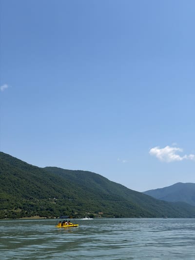 The image depicts a wide, tranquil body of water with lush, green mountains in the background, under a clear blue sky. In the foreground, a yellow pedal boat with a canopy is carrying four individuals. They appear to be enjoying a leisurely ride on the water. The water surface exhibits gentle ripples, reflecting the light and creating a serene atmosphere. Further in the distance, several small boats are visible on the water, and a few buildings are scattered along the shoreline, nestled amongst the trees. A jet ski is also visible in the distance, leaving a wake behind it. The overall scene conveys a sense of peaceful recreation in a natural setting. This location is likely on the Paliastomi Lake near the town of Avenisi in Georgia.
