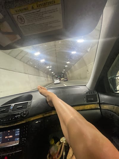 An interior view from a vehicle in Golemo Buchino, Bulgaria, shows a bare, tan-skinned leg and foot resting horizontally on the black dashboard. The foot is positioned above the central air vents, with the leg extending towards the lower right of the frame. The car's dashboard features silver-colored air vent grilles and control dials. Below the dashboard, a media system display shows "99.1 MHz" and menu options including "Bass," "Treble," "Bal/Fad," and "Back." A sun visor is visible in the upper left, displaying "WARNING" and "AVVERTENZA" text, along with a circular symbol that is partially obscured but appears to be a warning graphic.
Through the windshield, a brightly lit tunnel is visible. The tunnel walls are a light beige or grey, constructed from horizontal concrete segments. Numerous rectangular light fixtures are mounted overhead, illuminating the tunnel. The windshield surface displays multiple small specks, possibly water droplets or dust. In the distance, other vehicles are present within the tunnel, including a white car in the right lane. A green light, likely a traffic signal, is discernible further ahead in the tunnel. The view also encompasses the car's right A-pillar.