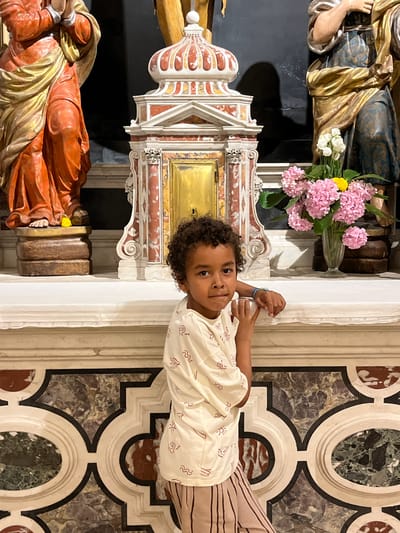 A young child with curly dark hair and brown eyes, wearing a cream-colored t-shirt with a red abstract pattern and striped pants, stands leaning against a white marble altar. The child is positioned to the right of the frame, looking towards the viewer. Behind the child, an ornate altar features a golden tabernacle, intricate red and white marble detailing, and two statues in religious attire, one on the left in orange and gold, and another on the right in blue and gold. A vase filled with pink hydrangeas and a single yellow flower sits on the altar to the right of the child. The altar rests on a patterned marble wall with circular and oval inlays. The scene appears to be inside a church or cathedral in Kotor, Montenegro.