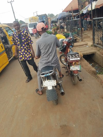 The scene depicts a busy urban street in Nnobi, Nigeria, under bright, overcast daylight.
In the foreground, a light brown dirt road occupies the lower portion of the image. On the left, an older male, dark-complexioned and bearded, wears a purple and yellow patterned long-sleeved shirt over a dark undershirt, dark trousers, and dark sandals. He holds a dark cane in his right hand and faces the camera, positioned next to a yellow commercial minibus with its door open, revealing a passenger's leg inside. To the right of the older male, a second male, dark-complexioned, is seen from the rear, seated on a silver motorcycle. He wears a light grey long-sleeved shirt, light grey trousers, dark flip-flops, and a reversed light pink baseball cap. The motorcycle has a dark blue seat, a white rear license plate, and an exposed exhaust pipe. Adjacent to this motorcycle, on the right side of the road, a second, red-brown colored motorcycle is parked, also facing away from the camera, with a green and white rear license plate and items secured to its rear rack.
The mid-ground reveals a concrete drainage ditch running along the right edge of the road, containing refuse and dark liquid. Behind this ditch, a row of market stalls with dark metal frames and various tarpaulin or umbrella coverings, including a prominent dark blue umbrella, display goods. Yellow sacks are visible near these stalls.
The background shows multiple pedestrians and vehicles, indicating a high level of activity. A white van is visible behind the seated motorcyclist, and further in the distance, a large blue truck is present. Buildings with reddish-brown roofs extend behind the market stalls. A tall utility pole with wires is visible on the far left against the bright sky.