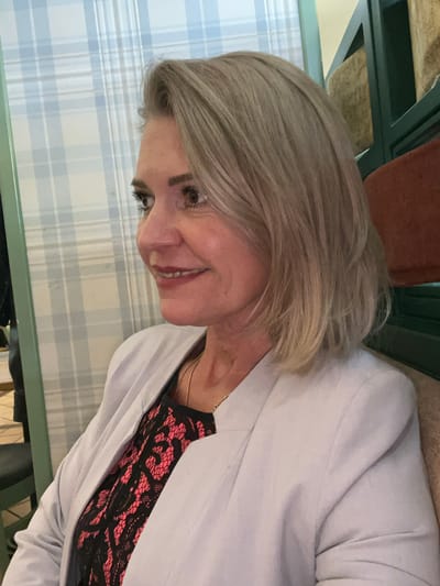 The image is a medium shot of a blonde woman in her 50s, turned to her left, looking slightly upwards. She has fair skin, light blonde hair cut to shoulder length with layers, and is wearing a light grey blazer over a black and pink patterned top. She has makeup on, including mascara and lipstick. Her necklace is visible. The background consists of a wall with a light blue and white plaid pattern wallpaper and a dark green shelf unit. The lighting suggests an indoor environment. There is no indication of the city of Kaliningrad, Russia, in the image.