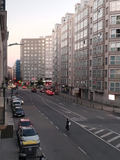 The image depicts a street scene in A Coruña, Spain, likely during the late afternoon or early evening given the sky's twilight hues and the streetlights beginning to illuminate. The perspective is from an elevated vantage point, looking down on a wide, multi-lane urban street lined with tall, modern buildings.
The buildings are contemporary in style, featuring multiple floors with numerous windows, many of which are bay windows that project outwards. The facades are predominantly light-colored, possibly stone or a light-colored render, and reflect the ambient light. Some windows show interior lights, suggesting occupancy.
The street itself is busy with vehicular traffic. Cars of various colors and models are visible, including a red car, a silver car, and a dark-colored SUV. Several cars are stopped at a traffic light, which is red in the foreground and appears to be red in the background as well. Other cars are in motion. Traffic signals, including red and green lights, are clearly visible.
On the sidewalk, in the foreground and to the left, several parked cars are visible. One car is a silver hatchback with a yellow bin positioned next to it. Further down the sidewalk, a lone woman with dark hair is walking away from the camera, carrying a white bag over her shoulder and a dark bag in her hand.
Further down the street, another pedestrian, a person wearing dark clothing and carrying a white backpack, is standing on the edge of the road, appearing to wait to cross. The road has clear lane markings, including solid white lines and dashed white lines, indicating traffic flow. There are also crosswalks painted on the road.
In the distance, more buildings and streetlights can be seen, contributing to the urban landscape. The overall impression is of a bustling city street experiencing the transition from daylight to evening.