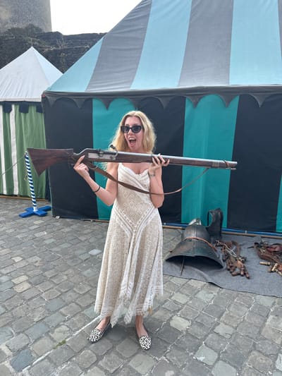 In Vianden, Luxembourg, a woman with blonde hair and sunglasses stands in front of a medieval-themed setup. She is wearing a white, lacy sundress and animal-print loafers. She holds a long, wooden rifle with a leather strap and appears to be shouting with excitement. Behind her, two tents are visible: one with green and white stripes and another with teal and black stripes. On the ground to the right, medieval armor and leather accessories are displayed on a grey cloth. The ground is paved with cobblestones.