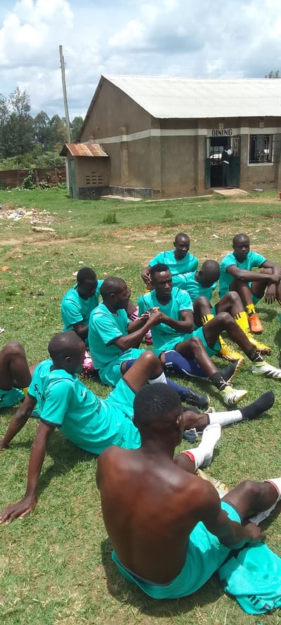 The image depicts a group of eight young men, all dressed in teal athletic shirts and shorts, sitting on a grassy field. They are all of African descent. The men are sitting in a circle, with their legs crossed and their arms resting on their knees. Some are wearing black athletic socks and shoes. They are seemingly relaxing and conversing with each other. There is a single-story building behind them with a sign that says "DINING" on it, and a telephone pole. The building is made of concrete and has a dirt roof. There are power lines overhead. The scene is most likely a recreational field or a schoolyard in Lubao, Kenya. The image is taken from a high angle. The day is sunny and the sky is partly cloudy.