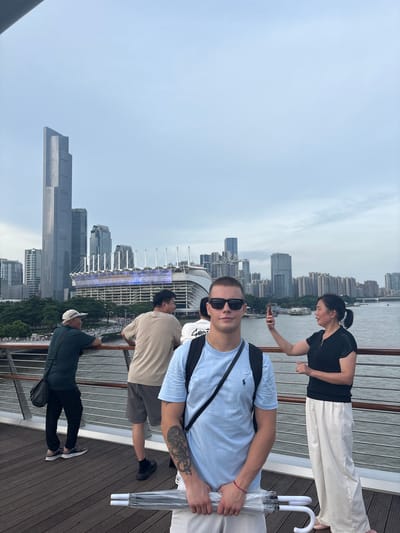 This is a medium shot of a group of people on a wooden boardwalk with a city skyline in the background. In the foreground, a young man with sunglasses and a light blue t-shirt stands looking at the camera, holding a clear umbrella. Behind him, a woman in a black shirt and white pants is taking a photo with her phone. Further back, two men are leaning on a railing, looking out at the water and the city. The skyline features tall modern buildings, including a prominent skyscraper, and a large stadium with a colorful illuminated facade. The sky is overcast. The location appears to be along a waterfront in Yuexiu District, China.