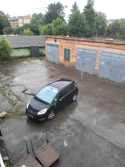 The image shows an outdoor scene in the city of Rivne, Ukraine, on a rainy day. The primary focus is a black Citroën C4 Picasso minivan parked on a wet asphalt surface. The minivan is facing towards the left of the frame, with its headlights on. Its windshield is covered in raindrops, and a blue sticker is visible on the upper right corner of the windshield. The license plate of the car reads "BK 4775 HO".
Behind the car, a row of brick garages with grey metal doors is visible. Two of these garage doors are numbered "21" and "20". To the left of the garages, there is a long, grey, corrugated metal building with a dark grey door. The ground is covered in puddles and reflections of the surrounding environment, indicating recent rainfall. Some patches of green grass are growing in the cracked asphalt. In the foreground, two metal carts with wire mesh baskets are present.
The background consists of trees and buildings, including a taller brick building with multiple windows and a construction crane visible above the treeline. The overall atmosphere of the image is overcast and damp, characteristic of a rainy day.