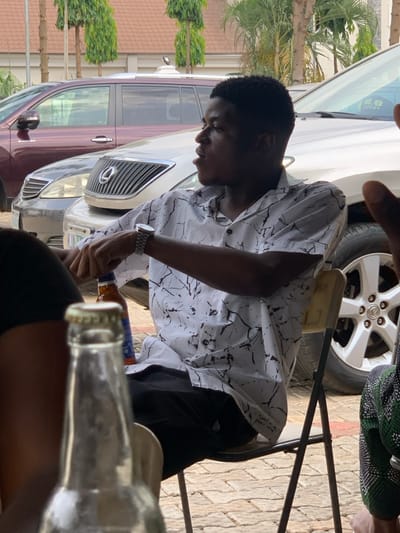 The scene is an outdoor setting, likely a paved parking area or driveway in Ogbunike, Nigeria. A young male, approximately 20-25 years old, is seated in the mid-ground, facing right with his mouth slightly ajar as if speaking. He possesses short, dark curly hair and wears a short-sleeved white collared shirt with black abstract patterns, dark trousers, a silver-toned wristwatch on his left wrist, and a silver-toned chain around his neck. His left hand holds an amber-colored glass bottle with a blue and white label. He is seated on a light-colored folding chair with dark metal legs.
In the extreme foreground, a blurred clear glass bottle with a gold-colored cap is visible. Partial views of other individuals are present: a dark-skinned arm/shoulder on the far left, and a person wearing a green, black, and white geometric patterned garment on the far right.
Behind the seated male, a light-colored SUV, identifiable by a Lexus grille and a partial "LAGOS" license plate, is parked. Further back, a darker, possibly maroon-colored SUV is also parked. The background features a building with a brown-orange tiled roof and several tall, green trees. The ground is covered with interlocking pavers.