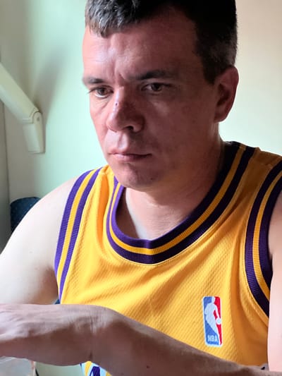 An adult male, appearing to be of Caucasian descent with short brown hair and a fair complexion, is captured in a close-up, medium shot. He is wearing a yellow basketball jersey with purple trim and an NBA logo. The man is looking downwards with a serious expression. He has a small mark or bump on his nose. His left arm is visible, resting in the foreground. The background consists of a plain, light-colored wall with a white medical-looking apparatus mounted on it. The lighting appears to be artificial and casts subtle shadows. The scene is indoors, and the overall impression is one of an informal or personal setting. There is no explicit indication of the location being Mozhga, Russia.