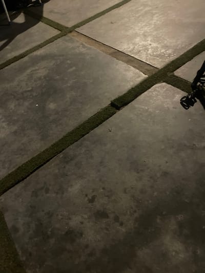 The image shows a concrete patio with artificial turf inlays, laid out in a cross pattern. The concrete is a dark gray color and appears to be slightly wet or stained in places. The artificial turf is a vibrant green and fills the gaps between the concrete slabs. There are some shadows on the patio, suggesting the presence of an overhead light source or perhaps natural light from a low angle. In the upper left corner, part of a dark object, possibly furniture or equipment, is visible, casting a shadow. In the upper right corner, a black object with some tangled cords is present, possibly an electrical cord or a piece of equipment. The scene is outdoors and appears to be nighttime or very early morning due to the lighting conditions. The location is identified as the city of Jimeta, Nigeria.