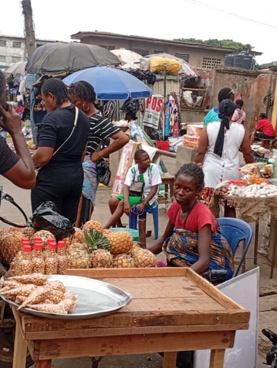 This image captures a vibrant and busy scene at an outdoor market, likely in an urban area in Africa. The overall lighting suggests it is daytime, possibly overcast, as there are no harsh shadows.

In the foreground, a wooden market stall occupies the center and left. On its flat surface, a silver metal tray holds numerous cone-shaped, light-colored snacks, possibly a local pastry or roasted nuts. To the right of the tray, several small glass bottles with red caps are filled with what appears to be groundnuts or peanuts. Behind these items, a large mound of fresh pineapples is piled up on the stall.

Seated behind this stall, to the right, is a young woman or older girl in a red t-shirt and a patterned blue and gold wrap skirt, resting on a blue plastic chair. She is looking directly at the camera with a neutral or slightly weary expression. Further back and to her left, another young person, a boy, wearing a white t-shirt with green accents (possibly a football jersey) and green shorts, sits on a blue plastic stool, looking away to the right.

The mid-ground is bustling with people and other market stalls. To the far left, the back of a person holding a phone to their ear is visible. Next to them, a woman in a black short-sleeved top and dark pants stands with her back to the camera. Behind her, another woman in a black and white striped top stands with her hands on her hips, partially obscured. Further back on the right, a woman in a white dress with a black head covering stands with her back to the viewer.

The background is filled with various market structures and buildings. Several large market umbrellas are open, providing shade, including a prominent blue one and a larger dark-colored tarpaulin or umbrella. Goods, including clothes and other merchandise, are visible on various stalls. On one stall in the background, a white sign with the bold red letters "POS" is clearly visible, likely indicating a Point of Sale or mobile money service. Smaller text below "POS" is present but largely unreadable, though "SALES" can be partially discerned. Behind the market activities, concrete buildings, some with corrugated iron roofs and a water tank, complete the urban backdrop.

The scene depicts the typical daily activity of buying and selling in a bustling outdoor market, characterized by numerous vendors, shoppers, and a variety of goods.
