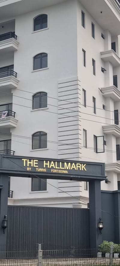 The image shows the exterior of a white, multi-story building, likely an apartment complex or hotel, in Moba, Nigeria. The building features balconies with black railings and numerous windows. A prominent sign above a dark gray gate reads "THE HALLMARK BY TURRIS FORTISSIMA" in illuminated gold lettering. The gate itself is made of vertical dark gray slats, and an outdoor lamp is mounted on a pillar to the right. The overall scene is clean and modern, suggesting a well-maintained property.