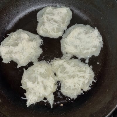 Four potato pancakes are being fried in a dark, cast-iron skillet with a black background. The pancakes are roughly circular and composed of shredded potatoes. They are submerged in hot oil, which is bubbling around them. The oil is a dark, glossy brown, with lighter oil pooling around the edges of the pancakes. The skillet's surface is also dark and slightly textured. The lighting is dim, suggesting an indoor setting, possibly a kitchen. The overall scene depicts the cooking process of potato pancakes. No people are visible. There is no visible text. The location context is Dunnae-myeon, South Korea.