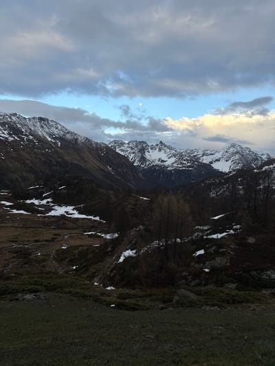 This photograph captures a rugged mountainous landscape in Poschiavo, Switzerland. The image is currently oriented vertically, with the sky occupying the left side and the ground the right. When viewed correctly in a horizontal orientation, the scene unfolds with a dramatic sky above snow-capped mountains and a grassy foreground.

The upper part of the scene (originally the left side) features a dynamic sky. On the far left, dark grey clouds are prominent, transitioning towards the center to a clearer blue sky where a distinct white, circular moon is visible. Further to the right, a band of clouds is illuminated with warm, golden light, indicating either sunrise or sunset.

The middle ground (central portion of the original vertical image) is dominated by a range of rugged mountain peaks and slopes. These mountains are largely covered with patches of snow, especially on their higher elevations. The lower slopes appear dark, covered in sparse, bare trees and rocky terrain, with additional scattered snow patches.

The foreground (originally the right side of the image) consists of uneven, dark green and brown grassy terrain, interspersed with rocks and darker patches of earth. No human activity, structures, or discernible text are visible. The overall lighting suggests either dawn or dusk, creating a subdued yet striking contrast across the alpine environment.