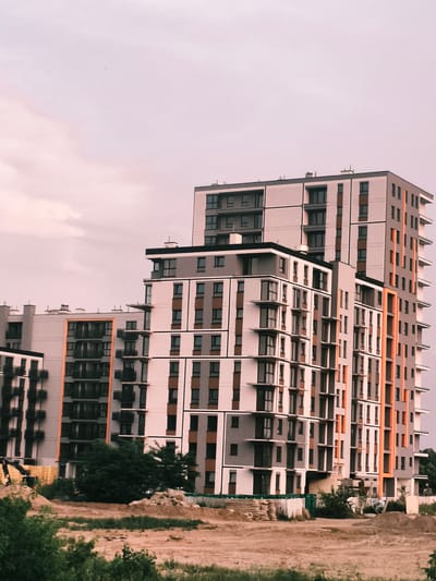 This image depicts a modern residential building complex under construction in Nowy Dwór Mazowiecki, Poland. The primary subjects are the multi-story buildings, characterized by their contemporary architectural design featuring a combination of light grey, white, and orange facades, with numerous windows and balconies. In the foreground, there is an area of disturbed earth, indicative of ongoing construction, with piles of soil and what appear to be construction materials such as wooden planks and possibly fencing. A yellow excavator is visible to the left, suggesting active construction work. Green trees and foliage frame the bottom and sides of the image, contrasting with the urban development.
The sky is a soft, muted pinkish-grey, suggesting either early morning or late evening light, possibly during twilight. The weather appears to be overcast, with subtle cloud formations visible. There are no people visible in the frame, and the focus is on the architecture and the construction site. The overall mood is one of development and progress, with the modern buildings standing starkly against the natural elements and the signs of construction. No visible text is present in the image.