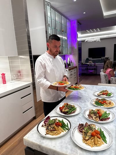 A male chef with short dark hair and a beard, wearing a white chef's jacket and dark pants, stands in a modern, high-end residential kitchen in Naples, United States of America. He is holding two dinner plates, one in each hand, and appears to be in the process of arranging or serving. On the light grey and white veined marble counter in front of him, seven additional identical plated meals are meticulously arranged. Each plate contains a main course consisting of a piece of grilled fish or poultry, a portion of orzo or grain pilaf, green beans, halved cherry tomatoes, small cooked carrots, a piece of radicchio, and a garnish of fresh herbs or microgreens.
The kitchen features white flat-panel cabinetry and a light-colored tiled backsplash. To the left, a red water bottle and a clear pitcher rest on the countertop. In the background, modern glass-front display cabinets with internal lighting, some exhibiting a purple hue, are visible, showcasing bottles and glassware. Further in the background, a living or dining area is partially visible, featuring a large wall-mounted television, light-colored seating arrangements, and a decorative chandelier. At least two individuals, including a person with blonde hair wearing a pink top, are seated in this distant area. The overall lighting is artificial and bright, with distinct purple accent lighting in the background.