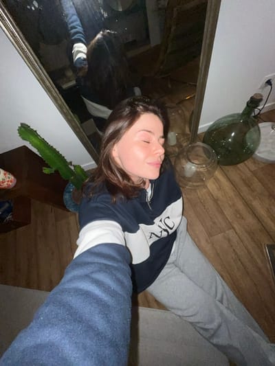This image, taken with a tilted perspective, captures a young woman resting indoors in Roubaix, France. She is lying on her back with her eyes closed, displaying a serene expression that suggests she is sleeping or deeply relaxed. Her dark brown hair is spread around her head. She is dressed casually in a navy blue and white zip-up hoodie, which features an indistinct white logo or text on the chest and white sections on the sleeves. She is also wearing light grey sweatpants. Her left arm extends into the foreground, possibly holding the camera for a selfie.

The setting is an interior space with warm wooden plank flooring. A large mirror with an ornate frame dominates the left side of the composition, reflecting parts of the room and the woman's silhouette. Beneath the mirror, a vibrant green, spiky cactus in a blue and white patterned pot adds a touch of nature. In the background, near the woman's head, decorative elements include a large, dark green glass demijohn bottle with a cork, and two clear glass vessels, one holding a pale object. The lighting is dim, suggesting an evening or nighttime atmosphere. The woman's legs are extended towards the right, resting on a grey, upholstered surface, likely a sofa. A portion of a patterned shoe is visible on the floor near the cactus. The image conveys a peaceful and private moment.