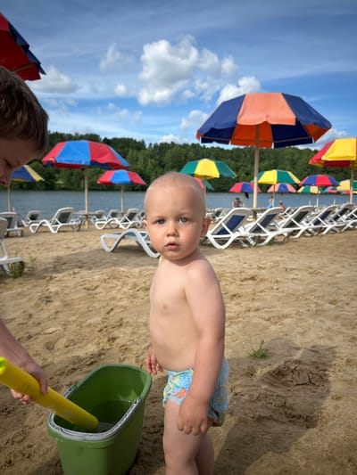 A young child, likely a toddler, stands on a sandy beach in the foreground of the image. The child has fair skin and light hair, with bright blue eyes looking directly at the camera. The child is wearing a diaper with a blue and orange pattern. To the left of the child, a person's arm, presumably belonging to an older child or adult, is visible, holding a yellow plastic shovel and a green bucket filled with water. The background of the image depicts a body of water, likely a lake, with a line of lounge chairs and colorful beach umbrellas visible behind it. Beyond the lake, a dense line of green trees forms the horizon under a partly cloudy sky. The overall scene suggests a recreational area, possibly a public beach within the city of Yubileyny, Russia.