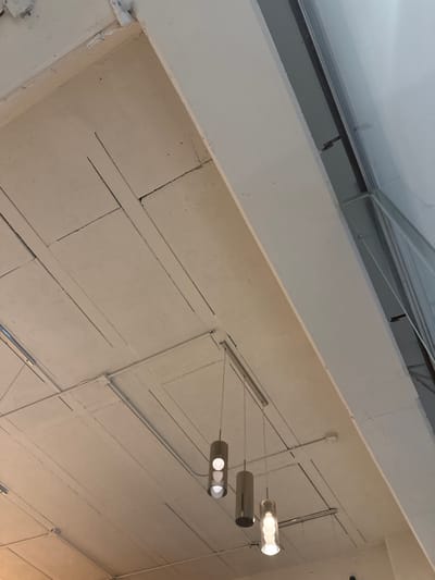 A low-angle shot captures a textured, white ceiling with visible grid patterns. A modern pendant light fixture with three cylindrical shades hangs from the ceiling, casting a soft glow. The architecture suggests an industrial or commercial space. Part of a skylight is visible at the upper left of the frame.