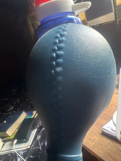The scene is depicted indoors in the city of Egan, Nigeria, likely on a desk or table surface. No individuals are present.
The primary subject is a large, dark blue, textured, bulbous object dominating the foreground and center. It features a prominent vertical line of raised, irregularly shaped bumps or nodules, reminiscent of a stitched seam or a grip pattern. The surface exhibits a finely stippled texture.
Behind and partially obscured by this blue object, a transparent blue bottle with a white and red flip-top cap is visible. To the left background, resting on a black and white marble-patterned surface, are several objects: a dark blue book, a book with a light-colored spine, and a black rectangular electronic device (possibly a power bank or smartphone) connected to a white charging cable. The cable extends downwards out of the frame. To the right background, a stack of white papers or cards rests on a light brown wooden surface. The lighting suggests an indoor environment with ambient illumination.
