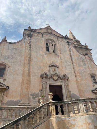 The image captures the facade of the Church of San Giuseppe in Taormina, Italy, viewed from a lower vantage point looking upwards. The church's architecture is Baroque, characterized by its weathered pinkish-beige stucco, ornate detailing, and a prominent central entryway.

Above the entrance, a niche contains a statue of a religious figure holding a cross. Flanking the entrance are decorative carvings and escutcheons. The building features arched windows on either side of the main facade. To the right, a slender, pointed steeple rises against the sky.

In the foreground, a stone balustrade and steps lead up to the church's entrance, suggesting a terraced approach. The balustrade itself shows signs of age, with visible weathering and discoloration. Two decorative urns, one with a small plant, are placed on either side of the staircase leading to the terrace.

The sky above is overcast with scattered clouds, casting a soft, diffused light on the scene. The overall impression is one of historical grandeur and a touch of decay, typical of ancient structures in southern Italy. No people are visible in the frame.