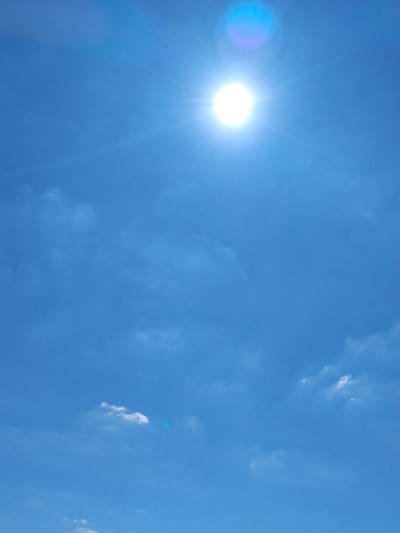 The image shows a bright sun in a clear blue sky with scattered white clouds. There is a lens flare effect from the sun, casting a bluish halo. The scene is an outdoor environment, characteristic of a daytime sky, and does not depict any specific urban features of Ustyvytsya, Ukraine. No people or objects are visible in the image.