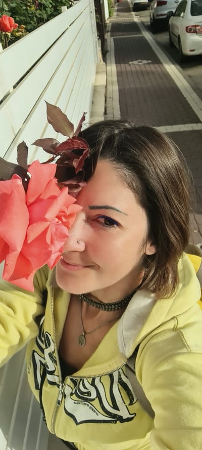 A selfie of a person with dark brown hair lying on a white slatted bench next to a large pink rose. The person is wearing a yellow zip-up hoodie with a black and white design on the front. They have a nose piercing and are wearing a dark beaded necklace and a silver necklace with a pendant. They have blue eyeliner. In the background, there is a street with cars parked along the side and buildings. Rose bushes grow nearby. This image could be located in Nahariyya, Israel, because the street with parked cars and the residential style buildings is commonplace in the city.