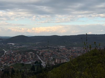 The image presents an elevated view of Bratsigovo, Bulgaria, under a cloudy sky. The town is densely populated with houses featuring red-tiled roofs. A main road cuts through the center of the town, with cars visible on its surface. Several multi-story apartment buildings are present. In the background, a line of low, forested hills stretches across the horizon. On the left, a winding road ascends toward an industrial-looking complex. In the foreground, the view is partially obscured by budding trees, indicating early spring. The overall color palette is muted, dominated by grays, greens, and browns.