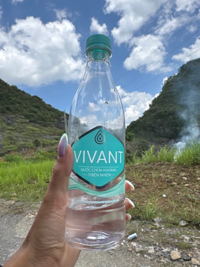A hand holding a partially empty, transparent plastic water bottle with a teal and white "VIVANT" label. The background shows a mountainous landscape with green vegetation, a blue sky with white clouds, and overhead power lines. A wisp of smoke is visible in the upper center of the image. The location is Lũng Cú Commune, Vietnam. A light-colored trail or road is visible in the foreground. The person has manicured nails with a light iridescent finish.
