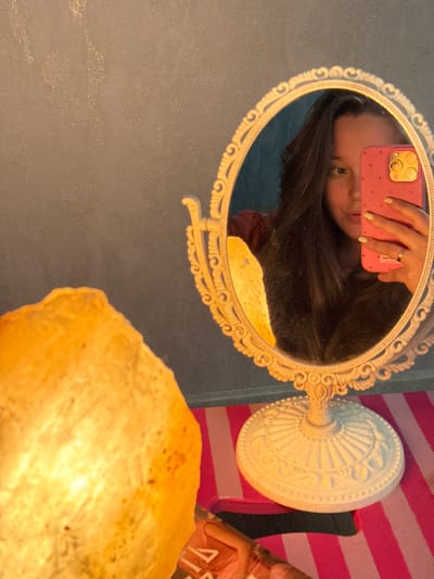 This media file is a selfie taken in a room. The main subject is a person taking the photo with their phone, whose reflection is visible in an ornate, oval mirror. The person is holding a pink smartphone with a dotted pattern and a raised camera module. Their left hand, adorned with glittery nail polish and a ring, is holding the phone. Their face is partially visible in the mirror, with dark hair falling around it. To the right of the mirror, a white ornate lamp base is visible, and behind it, a pink and white striped background suggests a decorative wall or fabric. The foreground is dominated by a large, illuminated object with a rough, crystalline texture, possibly a salt lamp. The overall lighting appears warm and ambient, suggesting it might be evening or indoors with artificial light. The setting seems to be a personal space, possibly a bedroom or dressing area. No text is clearly legible in the image.