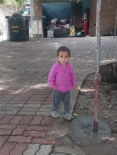 In Khakhnal, India, a young child stands on a patterned brick walkway. The child is wearing a vibrant pink knitted sweater and grey trousers, with grey clogs on their feet. Their hands are clasped in front of them, and they are looking directly at the camera with a neutral expression. To the child's left, a large black water tank with "DIPLASTIM" and "GOLD" labels is visible. Behind the water tank, a sign with text is posted. Further in the background, through an open structure with brick walls, a scene of cooking is apparent, with people preparing food over a fire and large pots. A red pole with white paint splatters stands to the right of the child, cemented into the ground. The environment appears to be outdoors, with dappled sunlight casting shadows on the walkway.