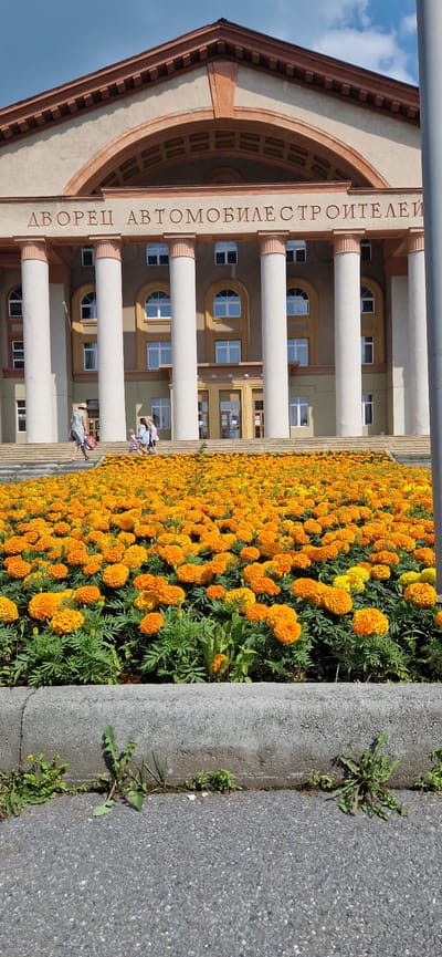 A low-angle, vertical, full outdoor shot shows the facade of a large, tan-colored building with a prominent arched entryway and tall, fluted white columns. The building has a pedimented roofline with decorative trim. Above the columns, a sign in Cyrillic letters reads "ДВОРЕЦ АВТОМОБИЛЕСТРОИТЕЛЕЙ" (Palace of Automobile Builders). In front of the building, a wide expanse of bright orange and yellow marigolds is in full bloom, separated from a paved area by a concrete curb. Three people, two women and a child, are visible on the stairs leading up to the building, and another woman walks down the stairs away from the camera. The sky is blue with scattered white clouds. This is likely the Palace of Automobile Builders in Miass, Russia.