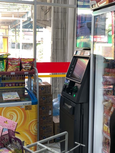 This image shows an interior view of a minimarket in Putat, Indonesia. In the foreground, a gray ATM machine is visible, displaying a green light indicating it is operational. To the right of the ATM, a glass-fronted display refrigerator is filled with packaged snacks. Behind the ATM, stacked cardboard boxes, possibly containing beverages, are visible. To the left of the ATM, shelves hold various packaged food items, including bags of "French Fries" potato chips. A large, yellow and orange swirl-patterned freezer is also present on the left side of the image. In the background, through a large window, an outdoor area with structures and vegetation can be seen. The scene suggests a typical retail environment. No people are clearly visible in the frame.