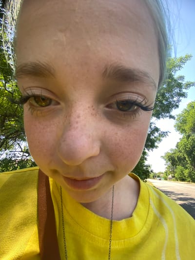 The image is a close-up, fisheye lens selfie of a young person outdoors. The subject has fair skin with freckles, light brown eyebrows, and noticeable false eyelashes. Their eyes are a greenish-brown. They are wearing a bright yellow t-shirt with white striped accents on the sleeve and a dark strap across their chest, likely from a bag. A thin, dark chain necklace is visible. In the background, lush green trees frame the scene, and a paved road with a faint tire track is visible leading into the distance, with a few houses and more trees in the far background under a clear blue sky. The overall setting suggests a rural or suburban environment. The location is identified as Nova Praha, Ukraine, implying the road and houses are in or around this town.