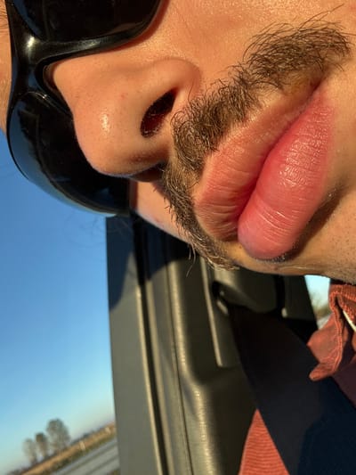 The image presents a close-up, angled view of a person's lower face, specifically the nose, mustache, and mouth, from a low-angle perspective. The individual possesses light-toned skin, with direct sunlight illuminating the right side of the face, highlighting pores on the nose. Black, reflective sunglasses partially obscure the upper left facial area. A dark, medium-length mustache is present, extending from beneath the nose and partially covering the upper lip. The lips are full, appearing hydrated, with the lower lip more prominent and slightly everted. Short stubble is visible below the lower lip and on the chin. The person wears a reddish-orange garment, likely a shirt, visible on the lower right. A dark, possibly grey or black, vehicle interior pillar or door frame runs vertically along the right side of the frame, casting a shadow. A dark strap, consistent with a vehicle seatbelt, is discernible diagonally across the garment. Beyond the vehicle interior, a clear, bright blue sky is visible. In the distant background, a flat, rural landscape featuring sparse trees and light-colored ground is seen, indicating an open area in the vicinity of Lodi Vecchio, Italy. The lighting suggests strong, natural sunlight, consistent with morning or late afternoon.