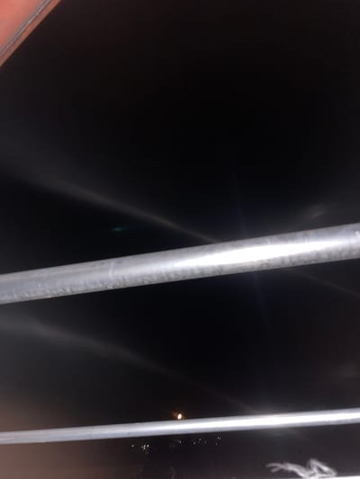 The media file shows a close-up of a dark scene, possibly at night, with two vertical metallic poles. The pole on the left is thicker and has a rougher texture, showing some discoloration. The pole on the right is thinner and appears smoother, with a bright white reflection running down its length, suggesting it's made of polished metal or is illuminated. The background is predominantly black, with faint, streaky light reflections that suggest a glossy or reflective surface, perhaps glass or metal. There are also some small, scattered points of light visible in the distance on the right side, which could be city lights or stars. A blurry, white, abstract shape is also faintly visible on the right pole, possibly graffiti or a natural smudge. The overall impression is of an urban or industrial setting at night, with artificial lighting creating reflections. There is no discernible activity or people, and the focus is on the textures and reflections of the metallic poles against the dark background. The color palette is dominated by black, grey, and white, with a hint of red on the lower left edge of the frame, likely from an object not fully in view.