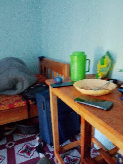 This image depicts an indoor scene within a room located in Sheywe ward, Kenya. The room features light blue walls and a floor with red, white, and dark patterned tiles.
To the left, a wooden bed frame supports a mattress covered with a red and gold patterned sheet. A large, gray, rolled-up blanket or duvet rests on the bed. A dark blue rolling suitcase stands vertically on the floor between the bed and a wooden table. Another dark bag is partially visible under the bed frame.
The central and right foreground is dominated by a wooden table. On the table, from left to right, are a small blue object, a dark-colored smartphone, a green plastic pitcher, and a yellow bowl containing residual food and a spoon. Another dark-colored smartphone lies on the table in front of the bowl. Behind the pitcher, a yellow and green plastic bag stands upright. No individuals are present in the scene.