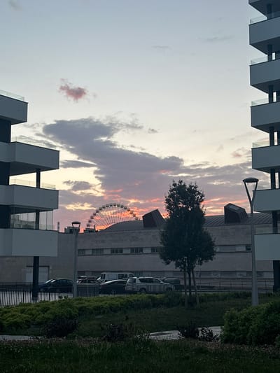 The image depicts a dusk scene in Gijón, Spain, with modern buildings flanking the foreground. A Ferris wheel is visible in the mid-ground, set against a sky painted with the warm hues of sunset. Clouds are scattered across the sky, some tinged with pink and orange. In the foreground, lush green bushes and grass line a paved area. A street with parked cars and a pedestrian walkway separates the foreground from a long, low-rise building with many windows. The architecture of the foreground buildings is characterized by sharp angles and balconies with glass railings. A street lamp stands on the right side of the image. The scene captures a serene urban landscape at twilight.