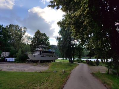 This media file captures a serene outdoor scene in Birštonas, Lithuania, likely during the late afternoon or early evening, given the soft lighting and the presence of dramatic, bright clouds against a blue sky, suggesting fair weather with some cloud buildup.
The primary subject is a large, grey boat resting on dry land, positioned on a concrete pad amidst lush green grass and mature trees. The boat appears to be out of the water, possibly undergoing maintenance or storage. It has a multi-level structure with railings and windows, suggesting it's a substantial vessel. To the left of the boat, a wooden structure and several large white bags are visible, hinting at ongoing work or supplies.
A paved pathway curves through the scene, leading towards the background where a body of water, likely a river or lake, is visible. The water reflects the sky and is bordered by trees and some man-made structures, possibly a park area with stone pillars and fencing. The surrounding trees are verdant and full of leaves, some with delicate, light-colored blossoms or seed pods hanging down, adding a touch of natural beauty to the scene.
The overall mood is peaceful and somewhat tranquil, with the natural elements of trees, sky, and water juxtaposed with the man-made object of the boat. There is no discernible activity or event taking place; the scene appears static, focusing on the environment and the beached vessel. No text is visible within the frame. The location cues, such as the style of the boat and the surrounding landscape, are consistent with a European setting.