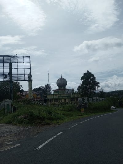 The image depicts a static daytime scene in Sandamal Eliya, Sri Lanka, featuring a mosque and its surroundings under a cloudy sky.
**Environment:** The sky is largely overcast with white and grey stratocumulus clouds. The foreground consists of a dark asphalt road curving from the lower right to the mid-left, marked with a solid white line and a dashed white line. To the immediate left of the road, an unkempt grassy area with various green plants and sparse vegetation is visible. Distant hills or elevated terrain, appearing vegetated, form the horizon in the background, partially obscured by cloud cover.
**Structures and Objects:**
* **Mosque:** A prominent mosque building occupies the midground. It is a light-colored structure, possibly light green or cream, with darker green trim and decorative arched elements. The main section features a large, ridged, metallic-toned dome, flanked by two smaller domes on the roof. Arabic script, including the word "MASJID" and other text, is visible above an entrance or central facade.
* **Minaret:** To the left of the main mosque building, a tall, slender minaret with a balcony and a smaller dome-like top stands. It appears to be light yellow or cream-colored.
* **Billboard Frame:** A large, dark-colored metal framework, consistent with an empty billboard structure, is positioned to the left of the minaret, partially obscuring other background elements.
* **Walls and Fencing:** A concrete wall, partially overgrown with vegetation, runs along the property boundary of the mosque. A small, yellow rectangular sign with red lettering is partially visible near this wall, adjacent to the road. A small, grey concrete structure, possibly a gatepost or utility box, is visible on the far left.
* **Vehicles:** A white passenger car is partially visible in the distance, on the curving road to the far right.
* **Vegetation:** Dense green trees and foliage are present around the mosque, behind the billboard, and on the distant hills.
**People, Actions, and Interactions:** No individuals are visible within the frame. No explicit actions or interactions are depicted; the scene is stationary.