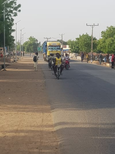 The image shows a street scene in Dabawa, Nigeria. The road is paved and runs horizontally through the frame. On the left side of the road, there are power lines and street lamps. Several people are gathered along the side of the road. Motorbikes and an IVECO truck are driving along the road. One man is walking on the side of the road. The sky is overcast, and there are trees lining the background.