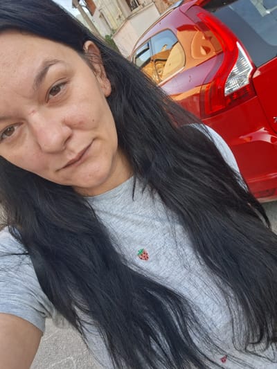 The image is a selfie taken outdoors in what appears to be a residential area in Gotse Delchev, Bulgaria. The foreground is dominated by a woman with long, dark hair. She is wearing a grey t-shirt with a small embroidered strawberry detail on the left side of her chest. Behind her, a bright red vehicle, possibly a Volvo, is parked. The vehicle's rear lights and part of its bodywork are visible, reflecting the surrounding buildings and sky. The background shows a building with balconies and windows, suggesting a street in the city. The overall lighting suggests it is daytime.