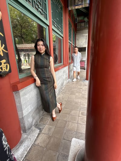 A woman in a dark, patterned, sleeveless dress stands by a window of a traditional Chinese building. She leans against the red-painted ledge of the building, her right arm resting on it. Her left leg is bent at the knee, with her foot in a brown pointed-toe shoe with a strap. A man in a light grey zip-up jacket and matching shorts walks away from the camera on a tiled path in the background. He is wearing white sneakers. The building features green-trimmed windows and red pillars. The architecture suggests a historical or cultural site, potentially within Xicheng District, China, known for its imperial palaces and traditional gardens.