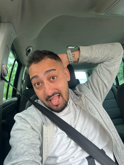 A selfie of a man in a car, angled such that the man's head is at the bottom of the frame. The man has brown eyes, short dark hair, and a trimmed beard. His tongue is sticking out. He is wearing a white shirt, a light gray zip-up hoodie, and a gray smartwatch with a dark-colored tattoo on the wrist. A black seatbelt crosses diagonally across his body. The car interior is visible, including the roof, dashboard, and window showing greenery outside. The location cannot be determined to be in Касапите, Bulgaria based on visual data.