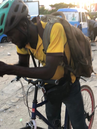 A man is on a bicycle in Ilado, Nigeria. He is wearing a yellow shirt with brown sleeves, black pants, a large brown backpack, and a gray and green helmet. The bicycle is blue. The location is a city street.